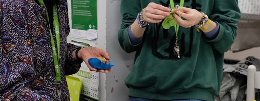 Two people wearing green lanyards stand at a table with mugs and plates of doughnuts. A pink tin of doughnuts is on the table, with bins underneath and posters in the background.