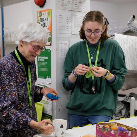 Two people wearing green lanyards stand at a table with mugs and plates of doughnuts. A pink tin of doughnuts is on the table, with bins underneath and posters in the background.