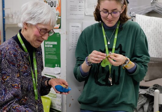 Two people wearing green lanyards stand at a table with mugs and plates of doughnuts. A pink tin of doughnuts is on the table, with bins underneath and posters in the background.