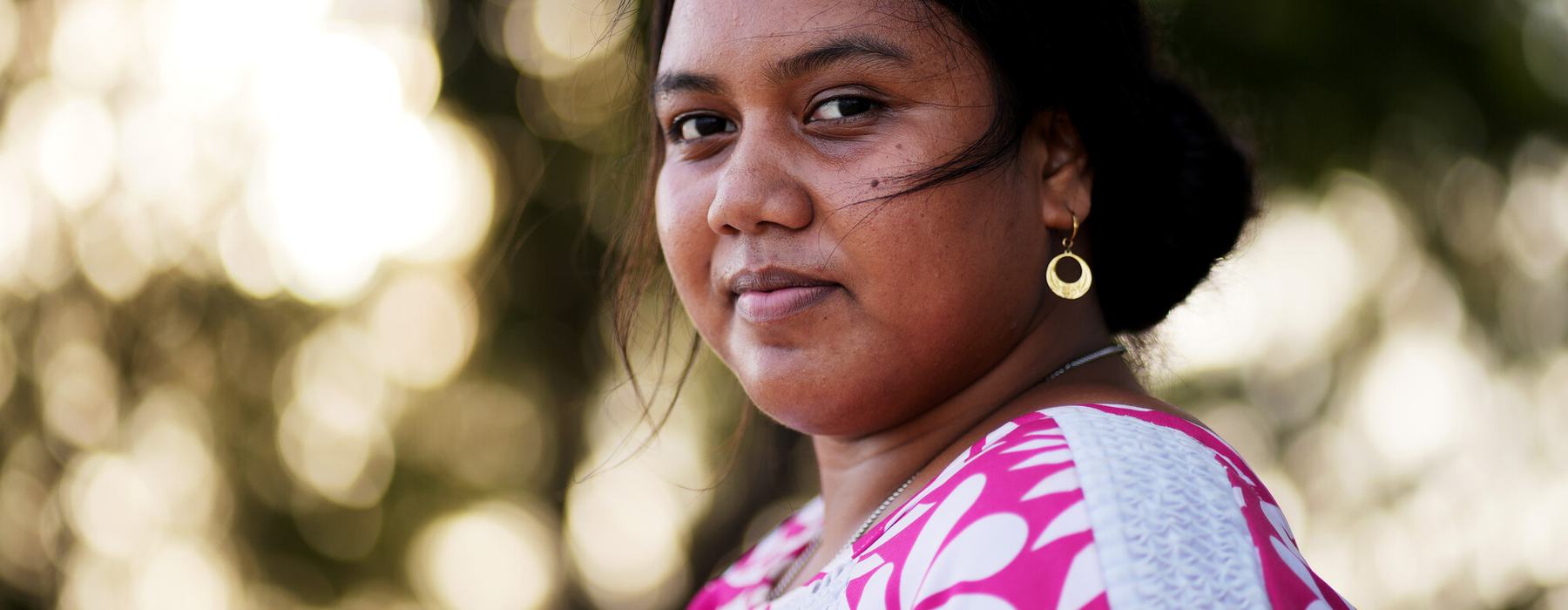 A dark-haired woman in a pink and white patterned top smiles at the camera with blurred trees in the background