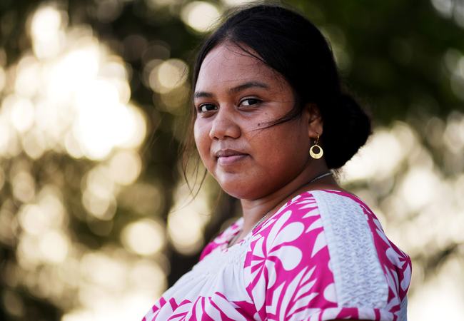 A dark-haired woman in a pink and white patterned top smiles at the camera with blurred trees in the background