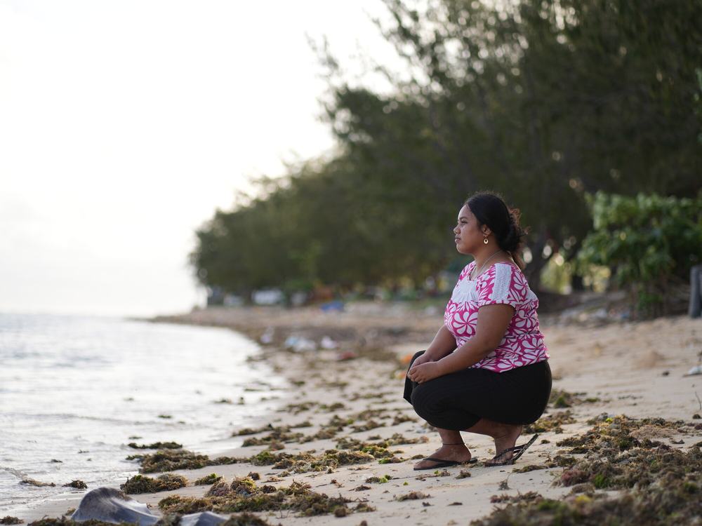 A woman crouching on a sandy beach by the water’s edge, looking out to sea.