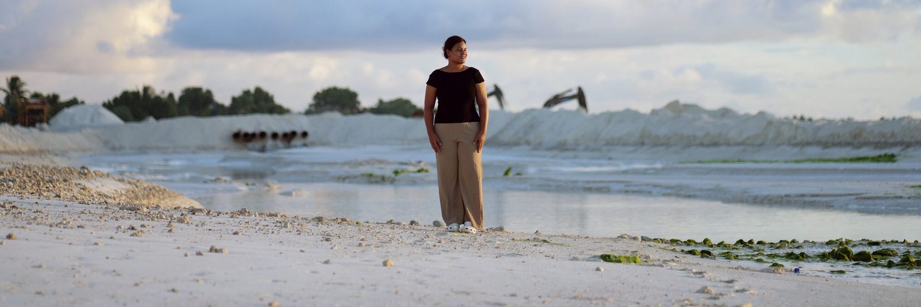 A woman wearing a black top and beige trousers is stood on a white sandy beach on the island of Tuvalu looking out into the distance.