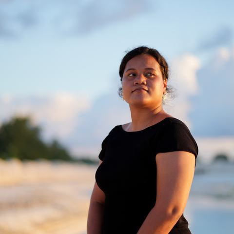 A woman in a black shirt stands by a calm seashore at sunset.