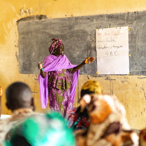 A community group is sat in a classroom in Adamawa State, Nigeria where a female dressed in a purple patterned garment is talking to the group