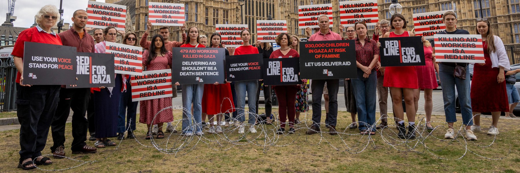 Protesters stand outside the UK Parliament holding signs demanding humanitarian aid access to Gaza, highlighting the risk of famine for 930,000 children. Placards read messages such as "Killing aid workers is a red line" and "Let us deliver aid."