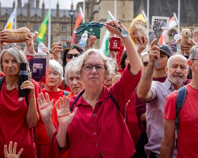 A group of Oxfam activists dressed in red hold symbolic items and raise their hands marked with red lines during a protest for Gaza. The demonstration takes place outside the UK Parliament.