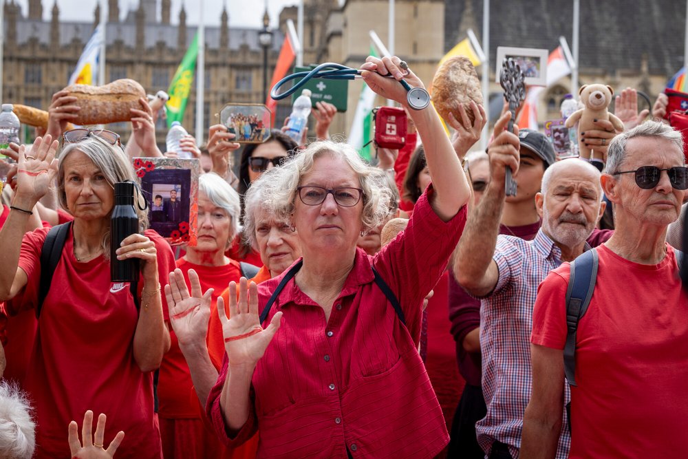 A group of Oxfam activists dressed in red hold symbolic items and raise their hands marked with red lines during a protest for Gaza. The demonstration takes place outside the UK Parliament.