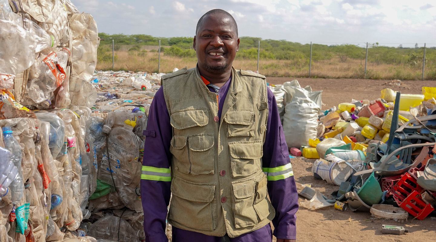 A smiling man in purple overalls and a tan-coloured utility waistcoat stands in next to a stack of compacted recycled material.