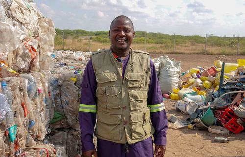 A smiling man in a khaki jacket stands next to a stack of compresses cubes of waste at a community waste centre in Mozambique.