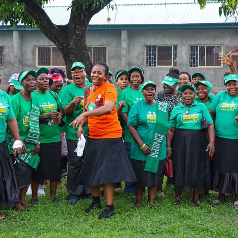 A woman wearing an orange t-shirt is stood in the middle of a large group of women wearing green t-shirts featuring the word 'Ogbonge'. They are all smiling and appear to be singing.