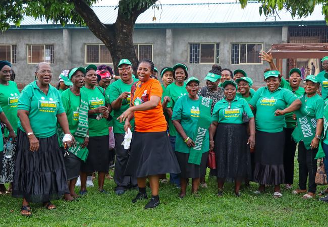 A woman wearing an orange t-shirt is stood in the middle of a large group of women wearing green t-shirts featuring the word 'Ogbonge'. They are all smiling and appear to be singing.