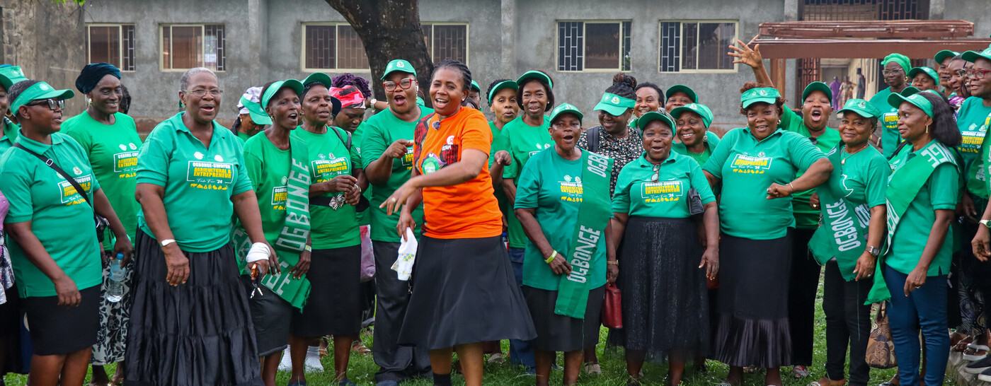 A woman wearing an orange t-shirt is stood in the middle of a large group of women wearing green t-shirts featuring the word 'Ogbonge'. They are all smiling and appear to be singing.
