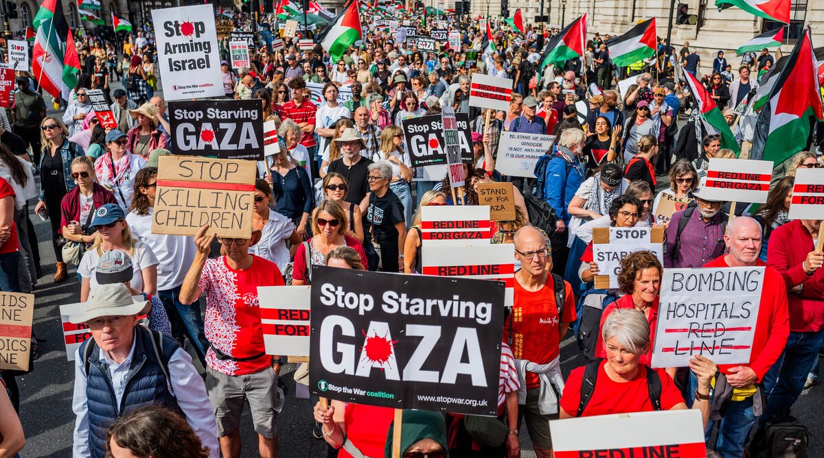 A large crowd of protesters march holding Palestinian flags and signs reading messages such as 'Stop Starving Gaza', 'Stop Killing Children', and 'Bombing Hospitals = Red Line'