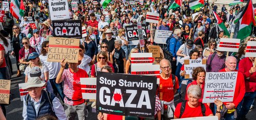 A large crowd of protesters march holding Palestinian flags and signs reading messages such as 'Stop Starving Gaza', 'Stop Killing Children', and 'Bombing Hospitals = Red Line'