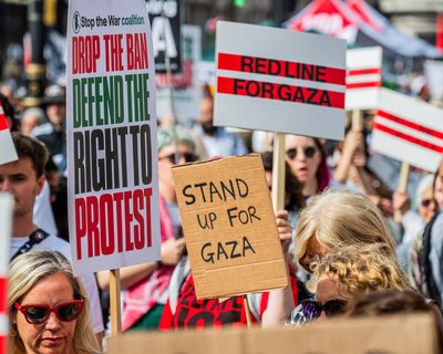 Protest scene with signs reading “DROP THE BAN DEFEND THE RIGHT TO PROTEST,” “STAND UP FOR GAZA,” and “RED LINE FOR GAZA.”