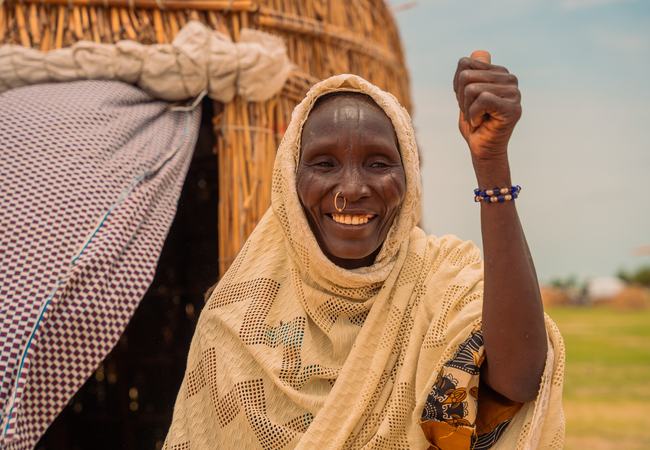 A smiling woman with a white head scarf and a nose ring stands outside her home in Lake Chad province.
