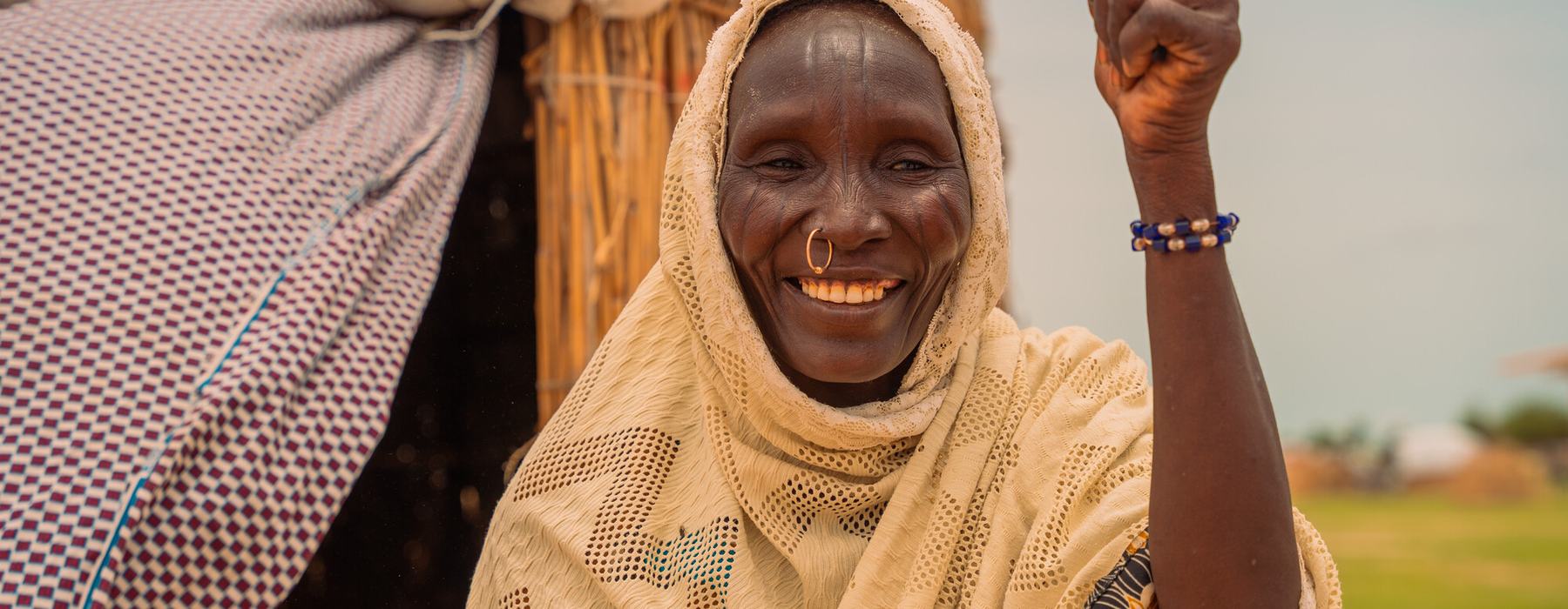 A smiling woman with a white head scarf and a nose ring stands outside her home in Lake Chad province.