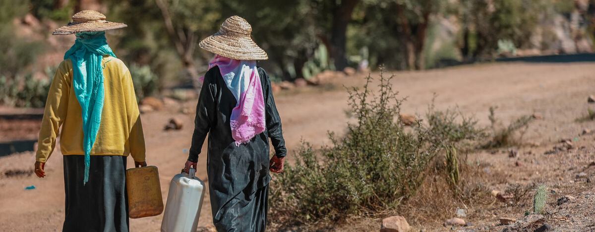Two people are walking along a dry track in Provincia de Taiz, Yemen, carrying large water containers.