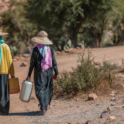 Two people are walking along a dry track in Provincia de Taiz, Yemen, carrying large water containers.