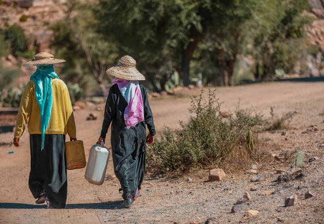 Two people are walking along a dry track in Provincia de Taiz, Yemen, carrying large water containers.