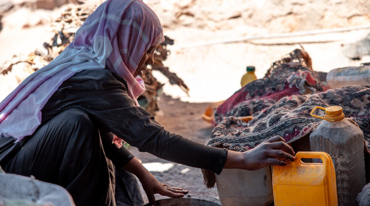 Abeer is crouching on the ground next to a tin plate while unscrewing the cap on a yellow water container.