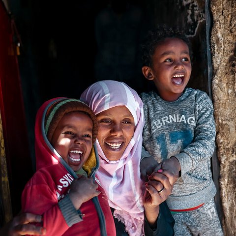 A woman and two children hold hands and smile in the doorway of their home in Taiz, Yemen, as the sun shines on them.