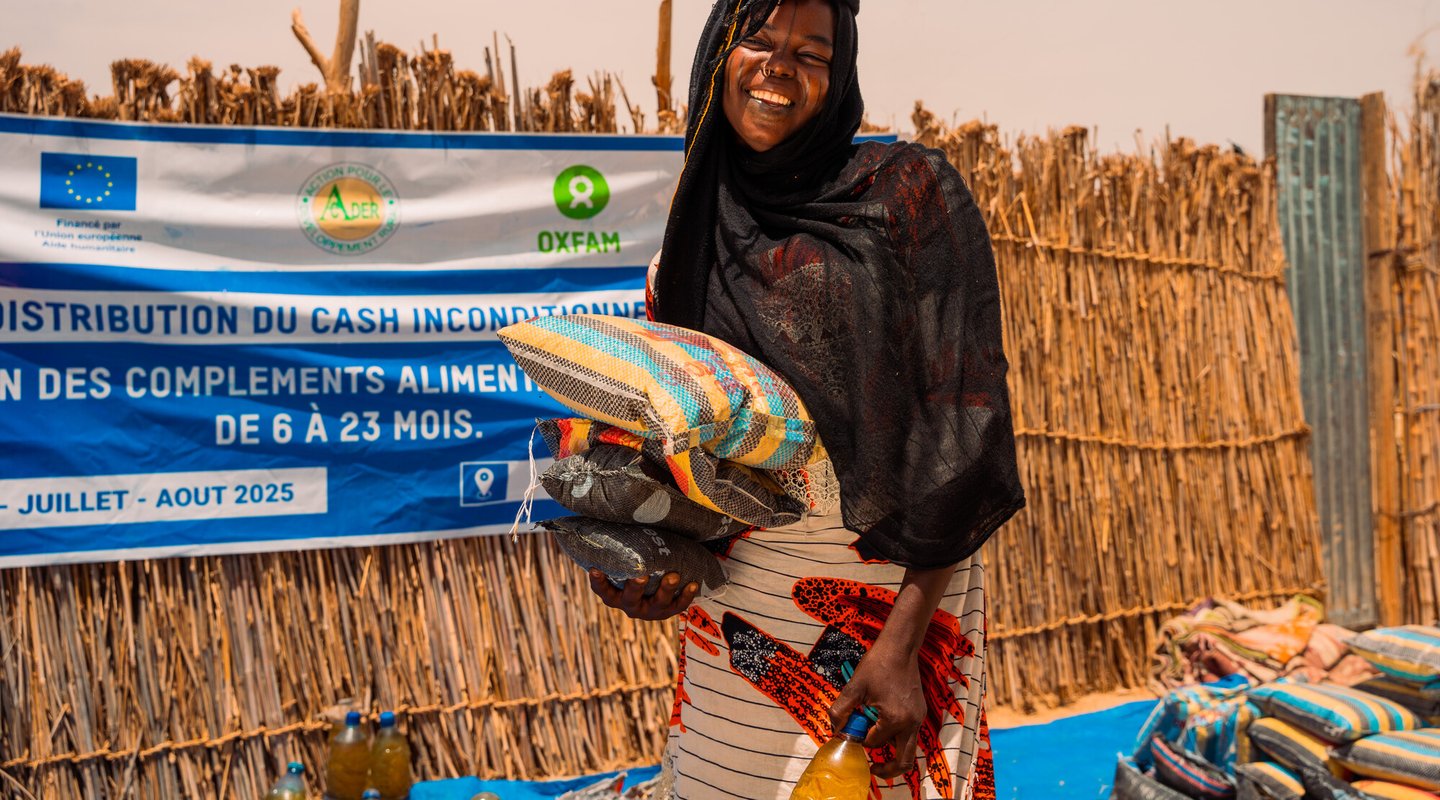 a woman holding sacks of food and smiling in front of a banner, which mentions cash and food supplement distribution.