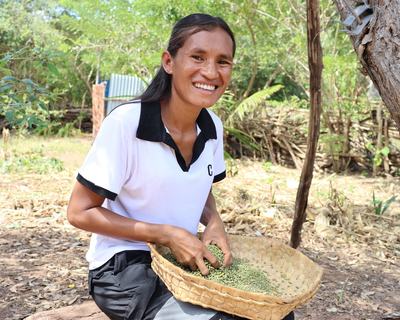 Person smiling into the camera, outdoors sorting grains in a woven basket beneath a tree in a rural setting.