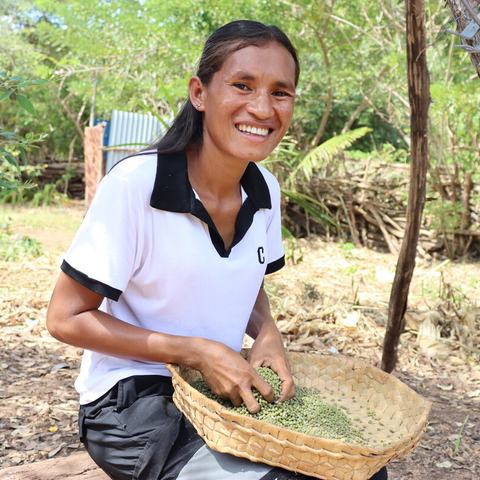 Person smiling into the camera, outdoors sorting grains in a woven basket beneath a tree in a rural setting.