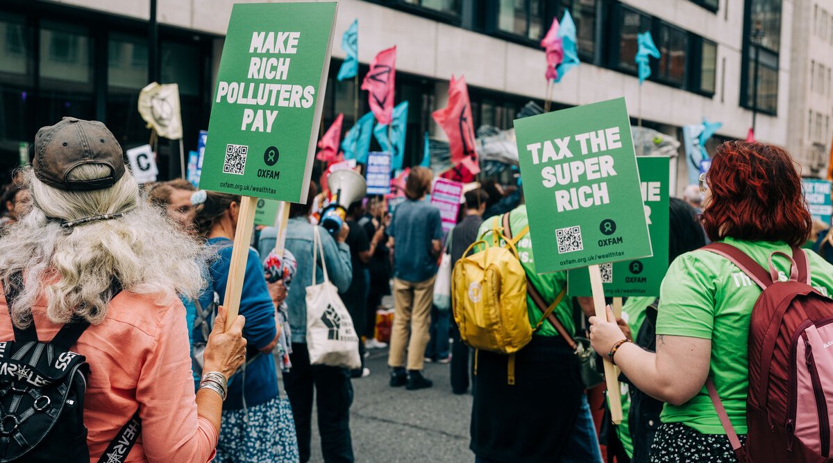 People taking part in a street protest holding green Oxfam signs that read “Make Rich Polluters Pay” and “Tax the Super Rich."