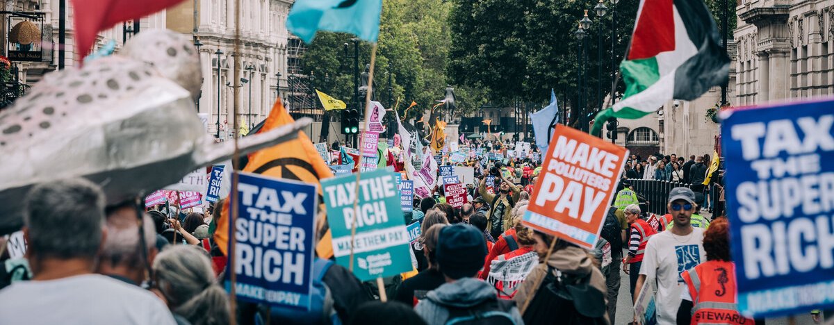 Demonstrators at the Make Them Pay march with Big Ben, London in the background. There are flags and banners with slogans of "Tax the super rich", "Tax the rich", and "Make polluters pay".