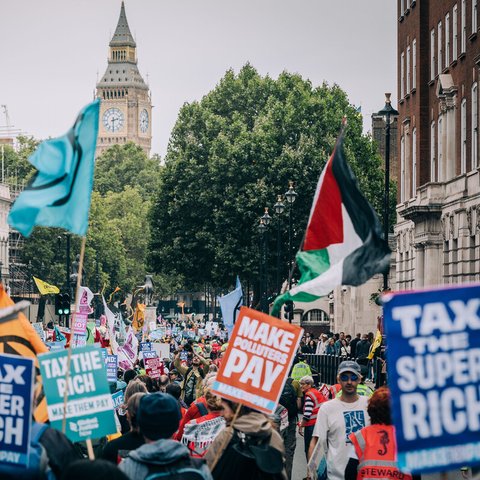 Demonstrators at the Make Them Pay march with Big Ben, London in the background. There are flags and banners with slogans of "Tax the super rich", "Tax the rich", and "Make polluters pay".