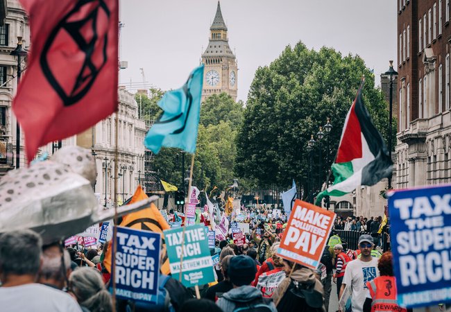 Demonstrators at the Make Them Pay march with Big Ben, London in the background. There are flags and banners with slogans of "Tax the super rich", "Tax the rich", and "Make polluters pay".