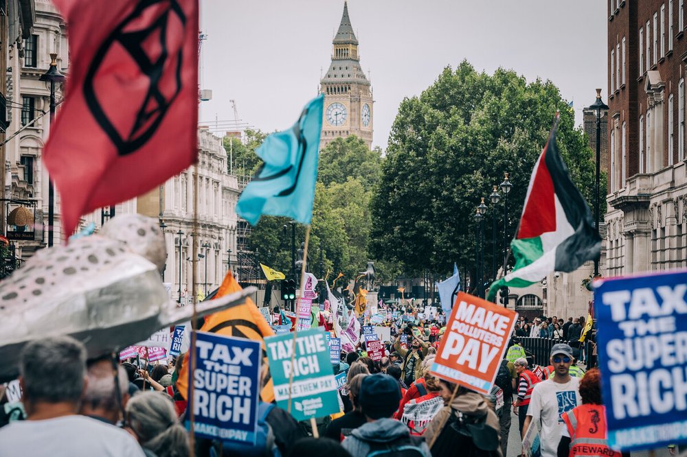 Demonstrators at the Make Them Pay march with Big Ben, London in the background. There are flags and banners with slogans of "Tax the super rich", "Tax the rich", and "Make polluters pay".