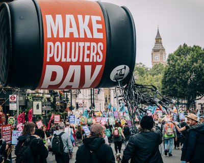A large protest march with colourful signs fills a city street, led by a giant inflatable oil barrel reading “Make Polluters Pay.” Big Ben is visible in the background as demonstrators walk forward together.