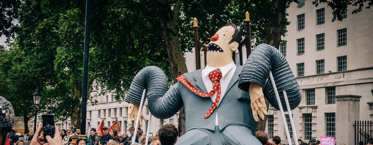 A large comical puppet of super-rich man in a grey suit sitting on a throne being held aloft by demonstrators at the Make Them Pay march.