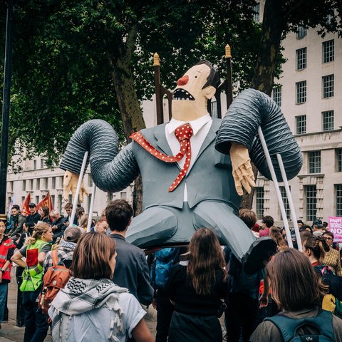 A large comical puppet of super-rich man in a grey suit sitting on a throne being held aloft by demonstrators at the Make Them Pay march.