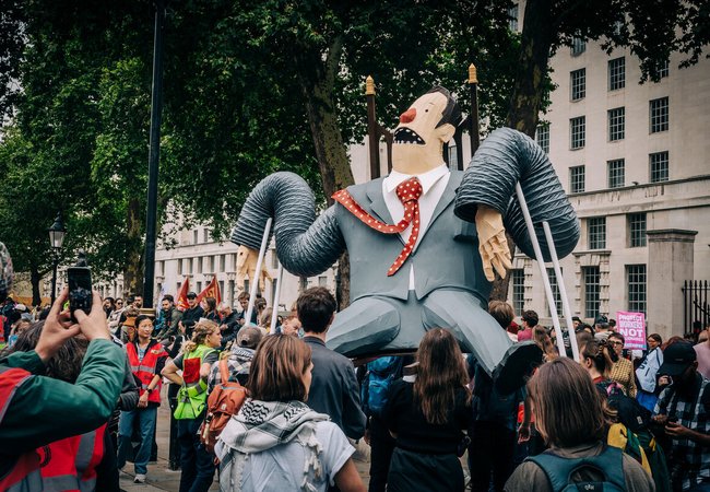 A large comical puppet of super-rich man in a grey suit sitting on a throne being held aloft by demonstrators at the Make Them Pay march.