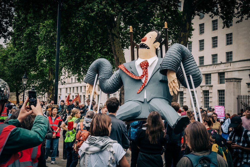 A large comical puppet of super-rich man in a grey suit sitting on a throne being held aloft by demonstrators at the Make Them Pay march.