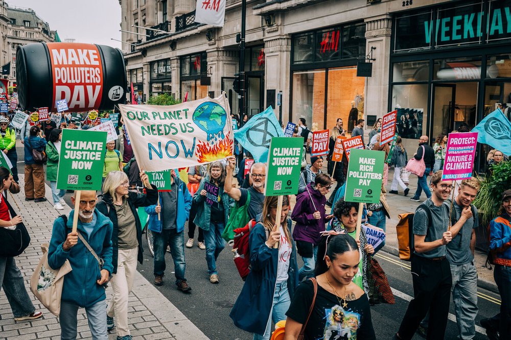 Protestors marching along a busy London shopping street, holding placards and banners. They read: "Climate justice now", "Make polluters pay" and "Our planet needs us NOW".