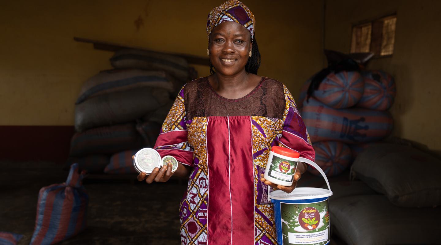 Safura, a smiling women in traditional Ghanan dress, holds up examples of the Shea Butter products she makes as part of her business.