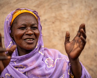 A smiling woman raising her hands as she speaks, dressed in a lilac embroidered shawl against a mud‑plastered wall.