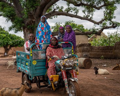 A group of women riding together in a motorised tricycle through their village, with goats and trees in the background.