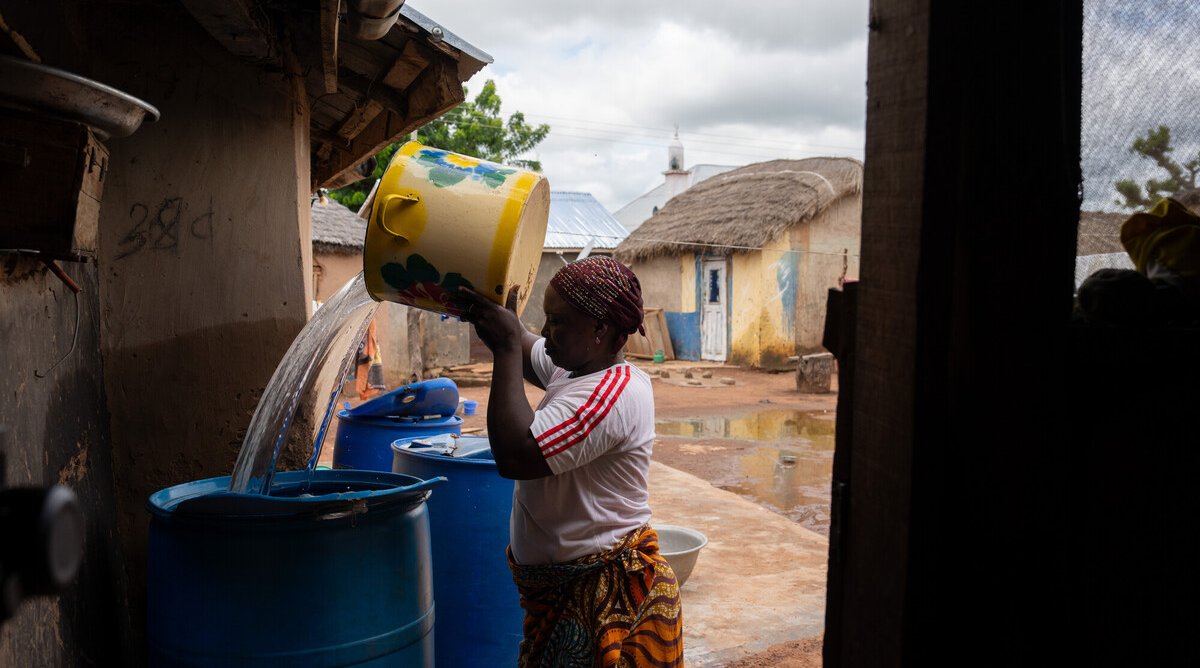 A woman pours water from a large yellow bucket into blue storage barrels in the yard outside her home, with traditional thatched buildings visible behind her.