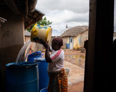 A woman pours water from a large yellow bucket into blue storage barrels in the yard outside her home, with traditional thatched buildings visible behind her.