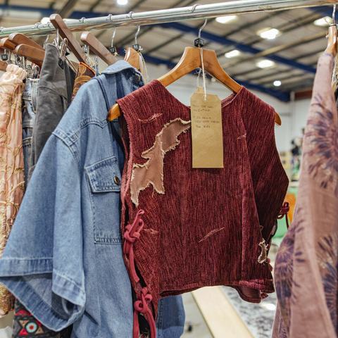 A clothing rail displaying a mix of second-hand garments, including denim shirts, patterned tops, and a red textured vest with visible patches and a handwritten tag attached.