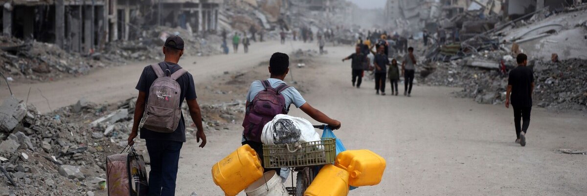 People walk through a street filled with rubble and destroyed buildings; one person pushes a bicycle loaded with yellow water containers and supplies. The scene depicts devastation and displacement.