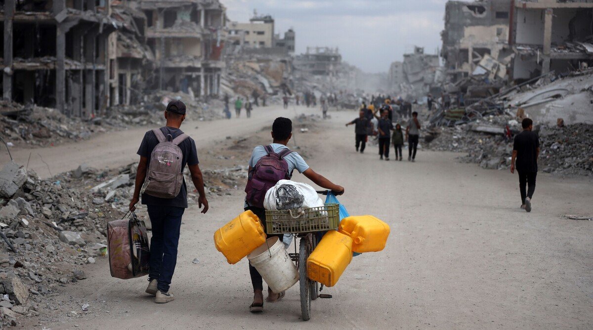 People walk through a street filled with rubble and destroyed buildings; one person pushes a bicycle loaded with yellow water containers and supplies. The scene depicts devastation and displacement.