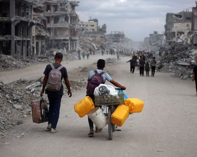 People walk through a street filled with rubble and destroyed buildings; one person pushes a bicycle loaded with yellow water containers and supplies. The scene depicts devastation and displacement.